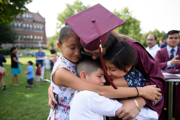 A newly graduated alumna, wearing maroon robes and mortarboard, stoops to embrace three children.
